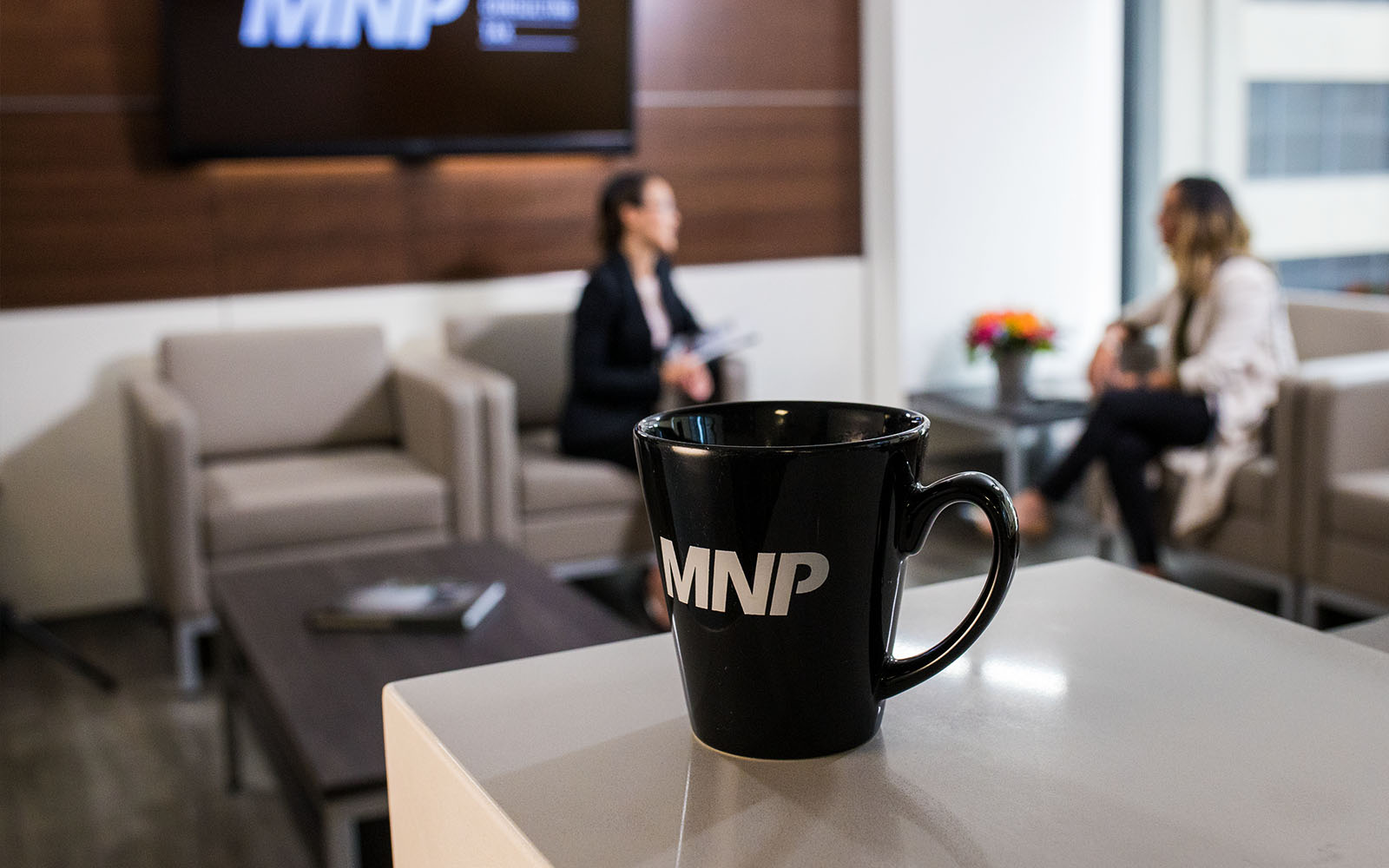 MNP mug placed on a desk with two people sitting in the background 