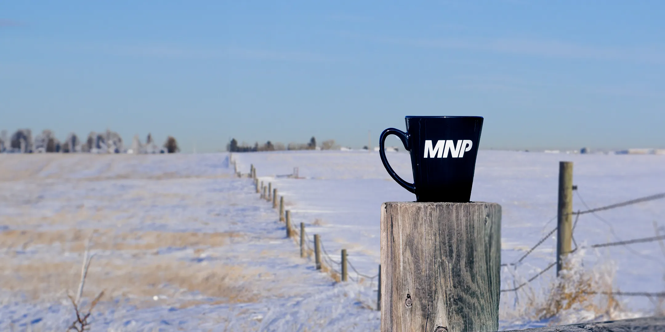 MNP mug sitting on a fence post with a winter farm land scene in the background.