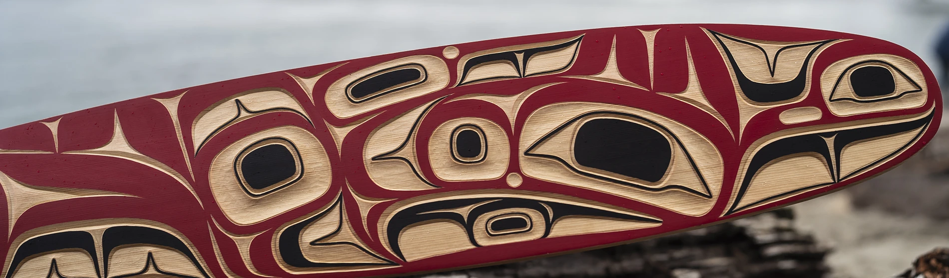 Close-up of a wooden paddle with Indigenous art in red, black, and natural tones.