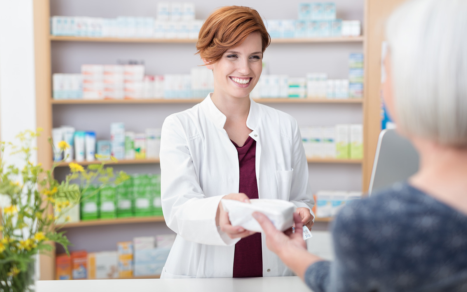 Smiling redhead pharmacist handing over prescribed medicines to an elderly female patient, view over the clients shoulder of the pharmacist