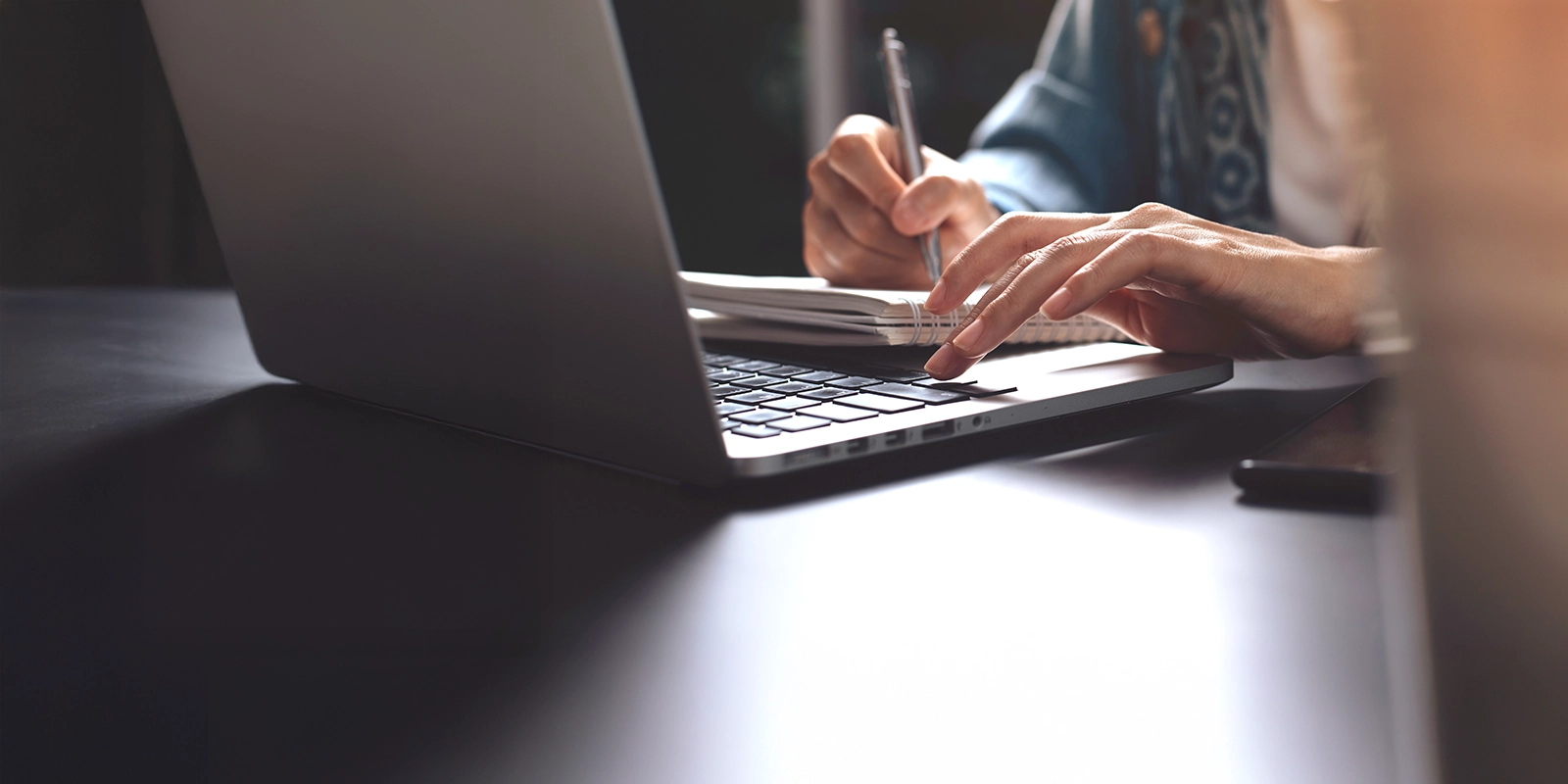 Person writing notes in a notebook while using a laptop.