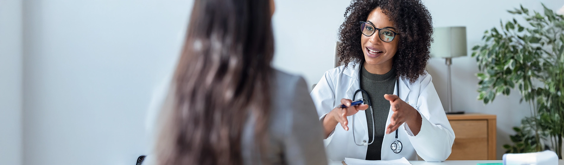 A healthcare professional in a white coat speaks with a patient across a desk, holding a pen and gesturing during the conversation.