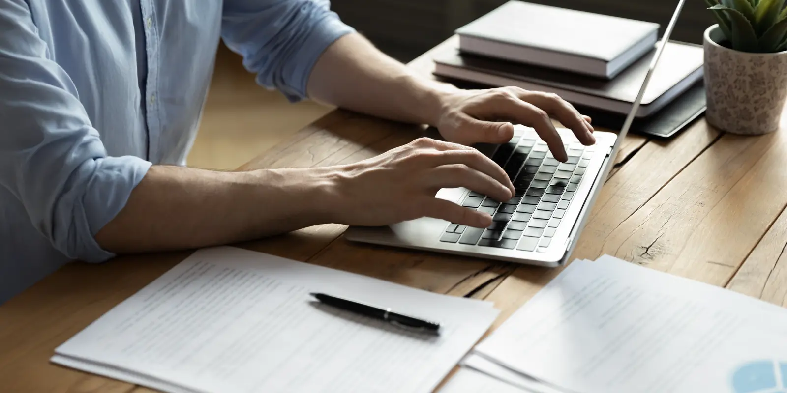 man typing on laptop while sitting at desk with documents and notebooks