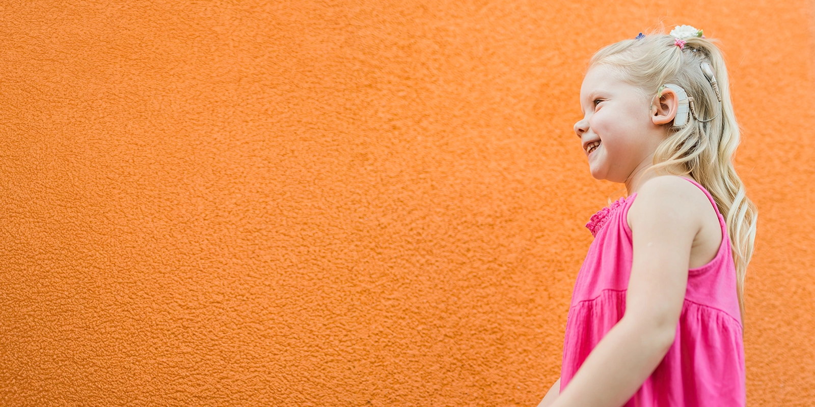 A young child with long blonde hair, wearing a pink sleeveless dress, is standing against an orange textured wall. The child's face is blurred for privacy.