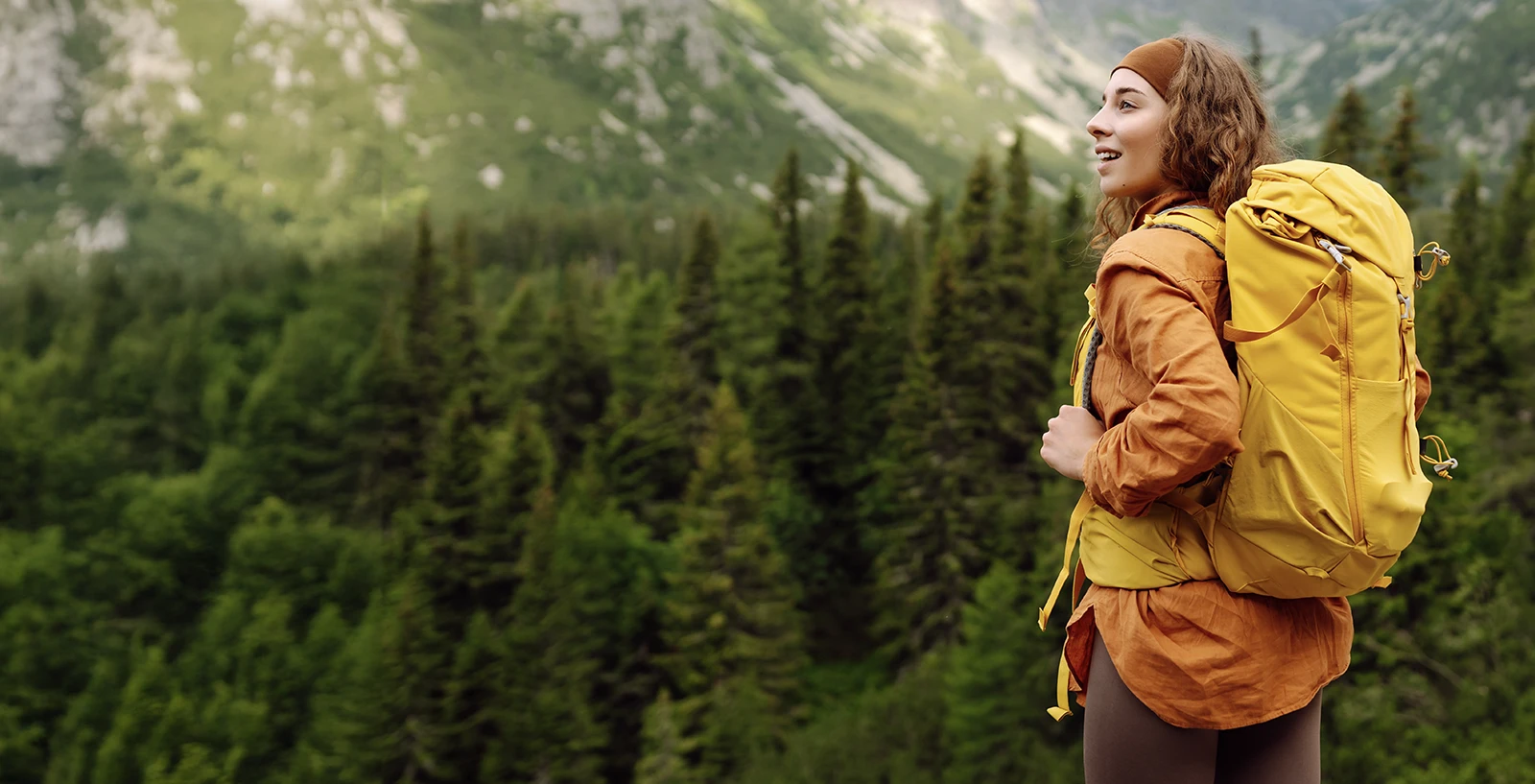 Woman wearing orange coat and yellow backpack walking through forest
