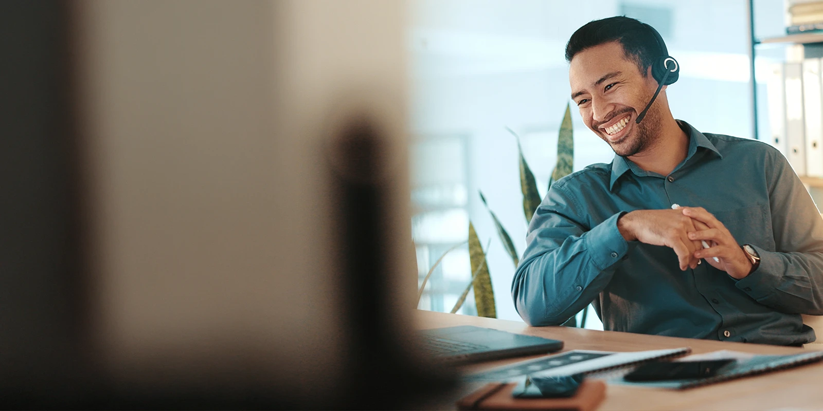 Person wearing a headset speaking at a desk with a laptop and smartphone, in a bright office setting with a plant in the background.