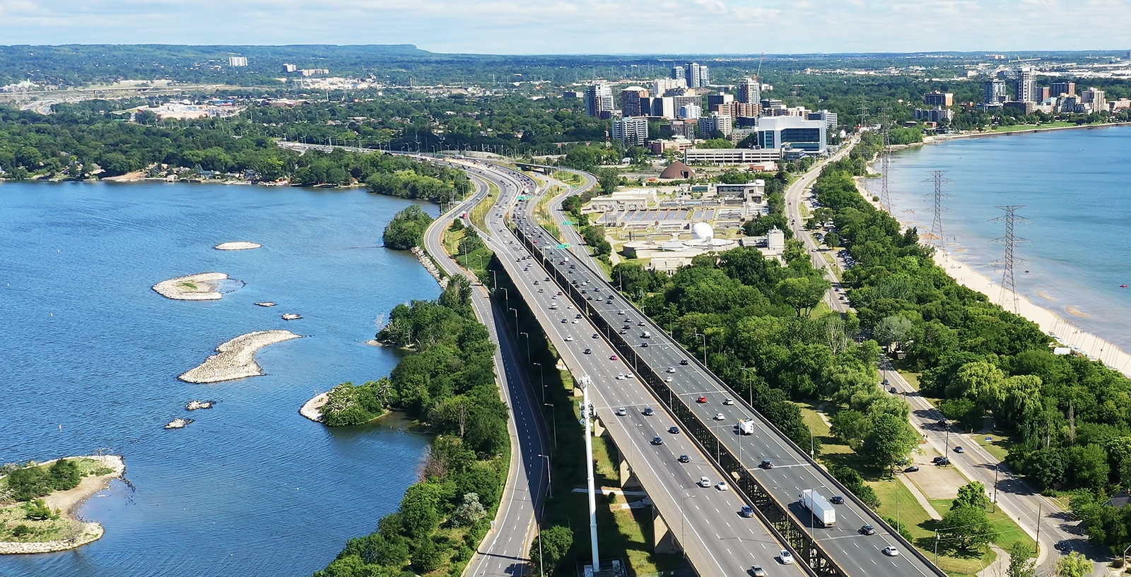 Aerial view of a highway next to a large body of water with small islands. The highway is bordered by greenery and trees, with a cityscape and buildings in the background. Power lines run parallel to the highway on the right.