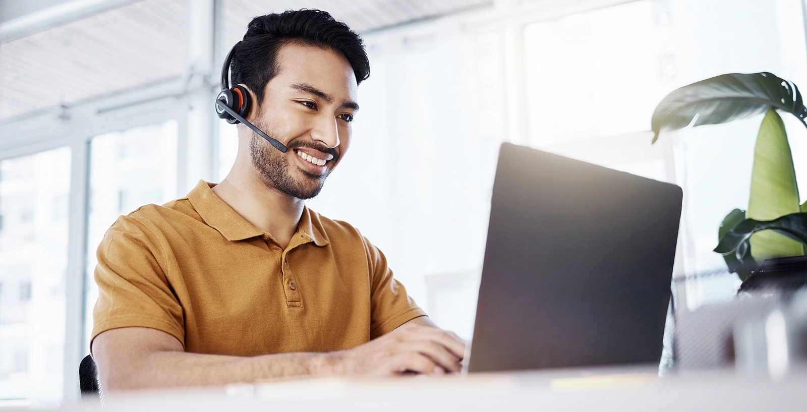A person wearing a mustard-colored polo shirt and a headset is sitting at a desk, working on a laptop.