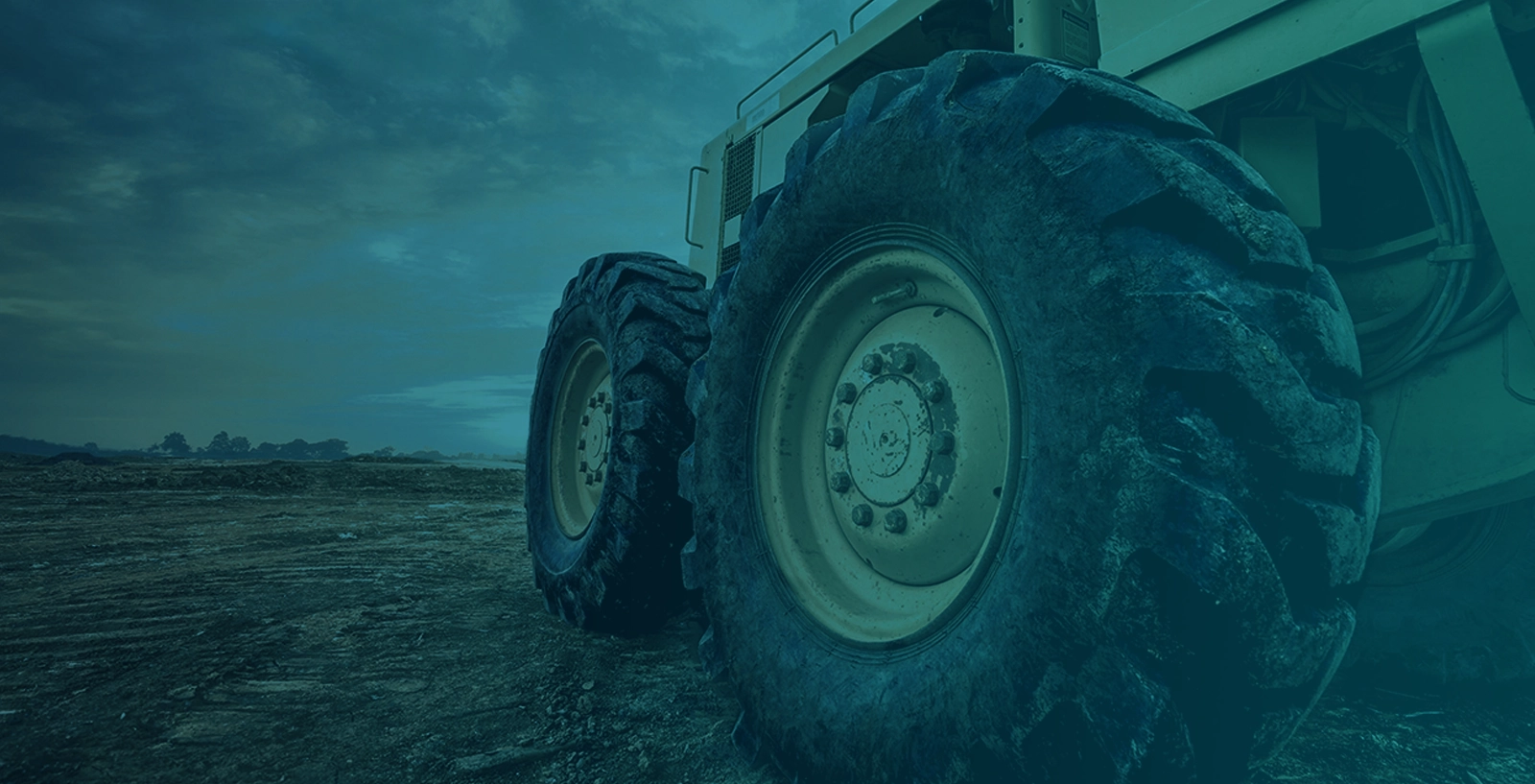 A close-up of large tires on heavy-duty equipment, positioned on rough terrain under a cloudy sky.