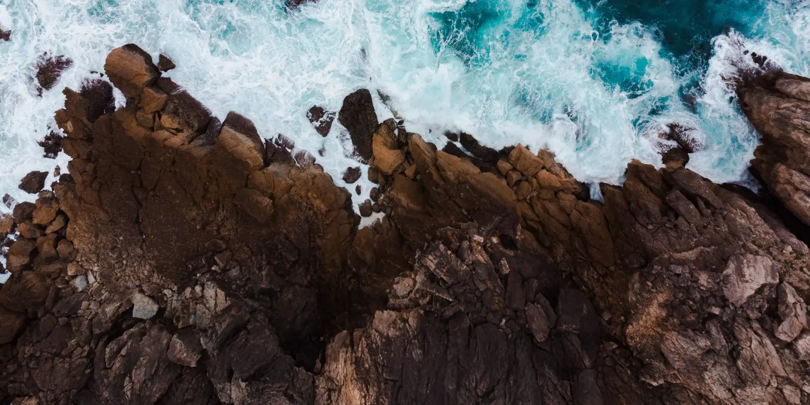 Vue aérienne d’un littoral accidenté avec des vagues s’écrasant contre des rochers dentelés. L’océan est d’un bleu profond avec de l’écume blanche.