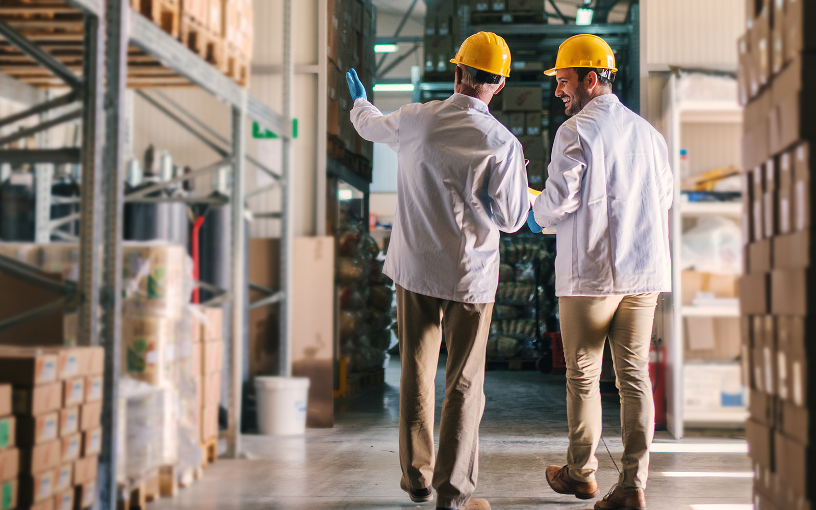 Father and son standing in their warehouse with helmets on their heads and looking at package prepared for transport