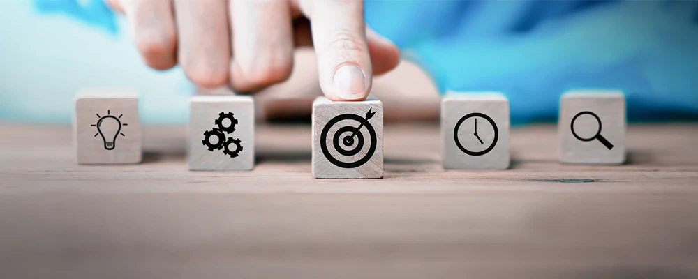 A hand points at a wooden block with a target icon, surrounded by blocks with lightbulb, gears, clock, and magnifying glass icons on a wooden surface.