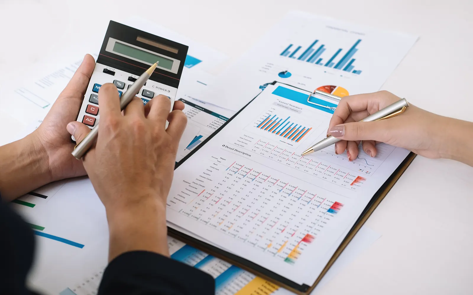 Close up of two people, one working on a calculator and another referencing data on a pile of paperwork 