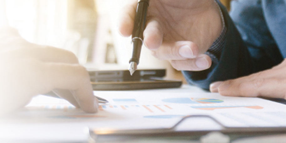 Two people look over papers with graphs and charts, one holds a fountain pen in his hand